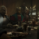 A group of reptilian aliens argue while sitting around a table with a lavish feast on it.